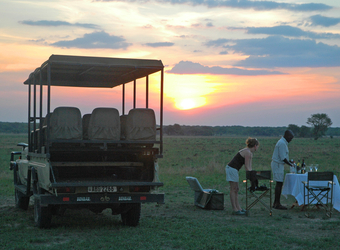 A safari guest prepares to eat in the secluded grasslands at a dining table with the gorgeous sunset and safari vehicle in Kafue National Park, Zambia