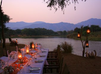 A gorgeous dining table is set out at twilight with lanters lighting the surrounding area with a view of the Zambezi River and mountains, Zambia