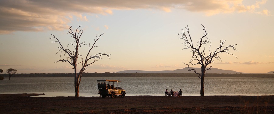 Safari guests eat a luxury picnic by Lake Malawi in southern Tanzania at sunset, a couple of crooked dead trees, safari vehichle, African safari 